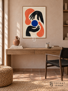 Graphic Hands by FREMAT in a light natural wood PVC frame with a flat white printed border. Creative maker study: pale clay-pink stucco wall, 150cm oak writing console, dark woven-cord chair at right, round rush ottoman left, warm quarry tile, diffuse morning light, one terracotta hand-built vessel.