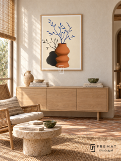Bright Vase by FREMAT in a light natural wood PVC frame with a flat white printed border. Ceramic artist sunroom: off-white rough limewash wall, 150cm blond ash cabinet, caramel rattan lounge chair left, low stone table, brick herringbone floor, bright morning light softened by reed blind and small green ceramic bowl.