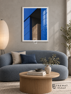 Blue Facade (PLC04) framed architectural wall art with a satin white PVC frame, flat white paper border and glass reflection. Calm apartment lounge, pale gray microcement wall, blue-gray curved sofa 180cm.