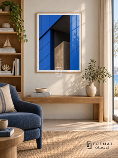 Blue Facade (PLC04) framed architectural wall art with a light wood-grain PVC frame, flat white paper border and glass reflection. Coastal library corner, warm white plaster, oak bookshelf to side.