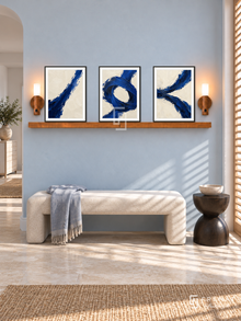 Ultramarine Tension complete shelf set in black frames: three 30 x 40 cm artworks with equal 2 cm borders, honey wood shelf and two sensor lamps. Coastal hallway in morning light.