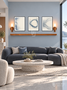 FREMAT Woven Water shelf set with three 30 x 40 cm black frames, 2 cm white borders, honey wood shelf and two warm sensor lamps. Morning contemporary waterfront lounge, mist blue wall, charcoal sculptural sofa, pale terrazzo oval table, pale grey limestone floor, sunny lake view window right.