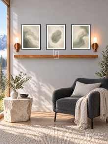 FREMAT Soft Topography shelf set with three 30 x 40 cm black frames, 2 cm white borders, honey wood shelf and two warm sensor lamps. Morning mountain modern reading area, fog grey plaster wall, charcoal upholstered armchair on black legs, irregular limestone side table, pale oak floor, sunny landscape window left.
