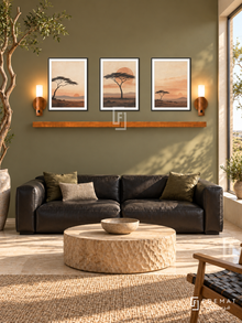 FREMAT Acacia Dusk shelf set with three 30 x 40 cm black frames, 2 cm white borders, honey wood shelf and two warm sensor lamps. Morning African contemporary lounge, muted olive wall, black leather low sofa, round carved pale timber table, cream stone floor, large sunny square window right, sculptural acacia branch at far left.