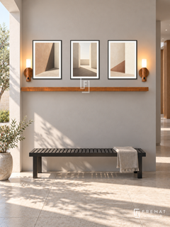 FREMAT Shadow Courtyard shelf set with three 30 x 40 cm black frames, 2 cm white borders, honey wood shelf and two warm sensor lamps. Morning architectural corridor, soft warm grey wall, black slatted bench, large pale stone tiles, sunny courtyard opening left casting geometric shadows, restrained contemporary decor.