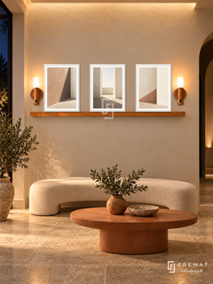 FREMAT Shadow Courtyard shelf set with three 30 x 40 cm white frames, 2 cm white borders, honey wood shelf and two warm sensor lamps. Night Mediterranean reception, warm ivory plaster wall, cream curved bench and terracotta oval table, limestone floor, dark arched doorway right, soft ceiling illumination.