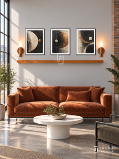 FREMAT Copper Orbit shelf set with three 30 x 40 cm black frames, 2 cm white borders, honey wood shelf and two warm sensor lamps. Morning industrial creative office, pale warm grey display wall, rust velvet sofa on black metal base, white round pedestal table, polished concrete floor, sunlit steel window left, brick column far right.