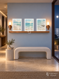 FREMAT Ocean Breath shelf set with three 30 x 40 cm white frames, 2 cm white borders, honey wood shelf and two warm sensor lamps. Night coastal entrance hall, pale blue wall, white curved bench, fluted stone side pedestal, limestone tiles, moonlit ocean door right, recessed warm ceiling.