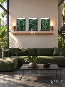 FREMAT Rainforest Layers shelf set with three 30 x 40 cm black frames, 2 cm white borders, honey wood shelf and two warm sensor lamps. Morning urban conservatory lounge, warm concrete wall, deep green modular sofa, black metal coffee table, dark slate floor, tropical garden window left, steel stair at far right.