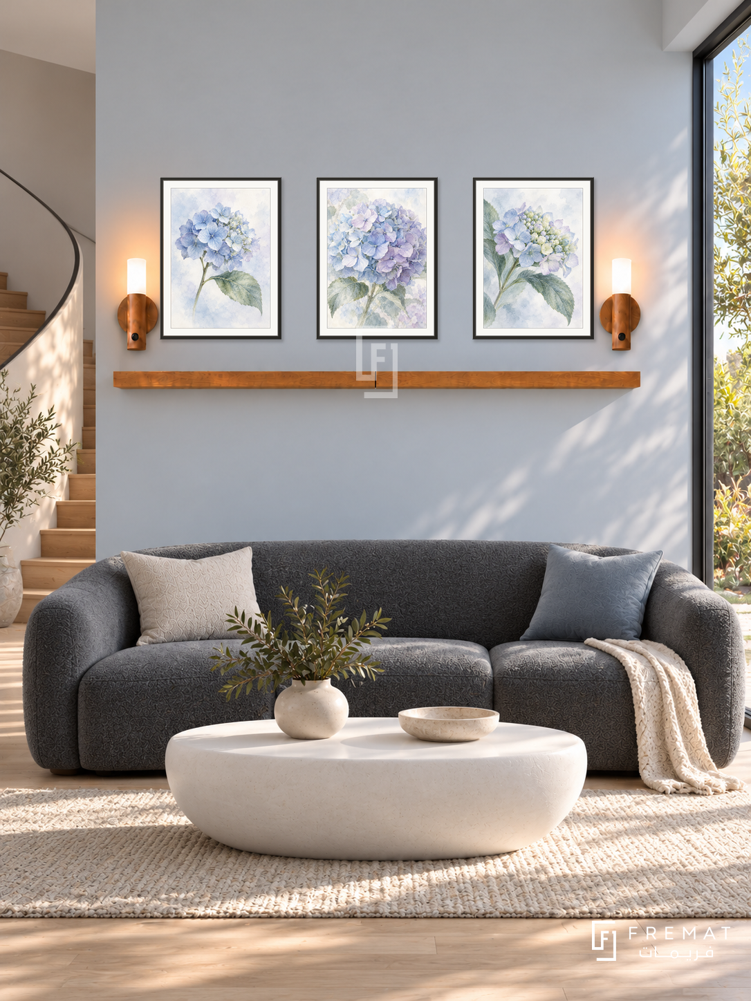 FREMAT Hydrangea Mist shelf set with three 30 x 40 cm black frames, 2 cm white borders, honey wood shelf and two warm sensor lamps. Morning soft contemporary living room, pale blue-grey wall, charcoal rounded sofa, white oval ceramic table, light timber floor, garden corner window right, curved staircase left.