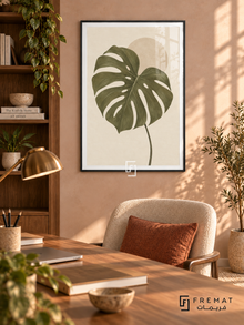 Monstera Study. An olive-green monstera leaf overlaps a pale sand disk on an ivory background. Shown in a Black PVC frame in a styled room.