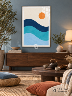 Blue Tide. Three flowing navy, turquoise and pale blue wave bands sit below an orange sun in an ivory sky. Shown in a Gold PVC frame in a styled room.