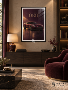 Aston Martin DB11 (car19), Black frame with white printed border in a styled interior. A burgundy Aston Martin DB11 sits on a glossy dark floor under a warm diagonal shaft of light. Gold-toned ASTON MARTIN DB11 lettering completes the wine-red composition.