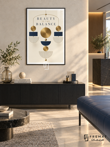 Beauty in the Balance (com20), Black frame with white printed border in a styled interior. A symmetrical hanging arrangement of gold discs, navy blocks and striped cream semicircles forms a geometric mobile. Widely spaced lettering reads “BEAUTY IN THE BALANCE” on ivory.