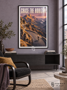 Chase The Horizon: A cyclist follows a winding mountain road through ochre peaks and lavender sky in a textured landscape illustration. Black frame with a white printed border, displayed in a styled room.