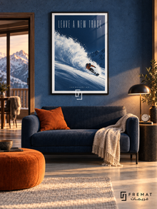 Leave A New Trace: A snowboarder in an orange jacket carves through powder on a deep cobalt slope beneath tall, light lettering. Black frame with a white printed border, displayed in a styled room.